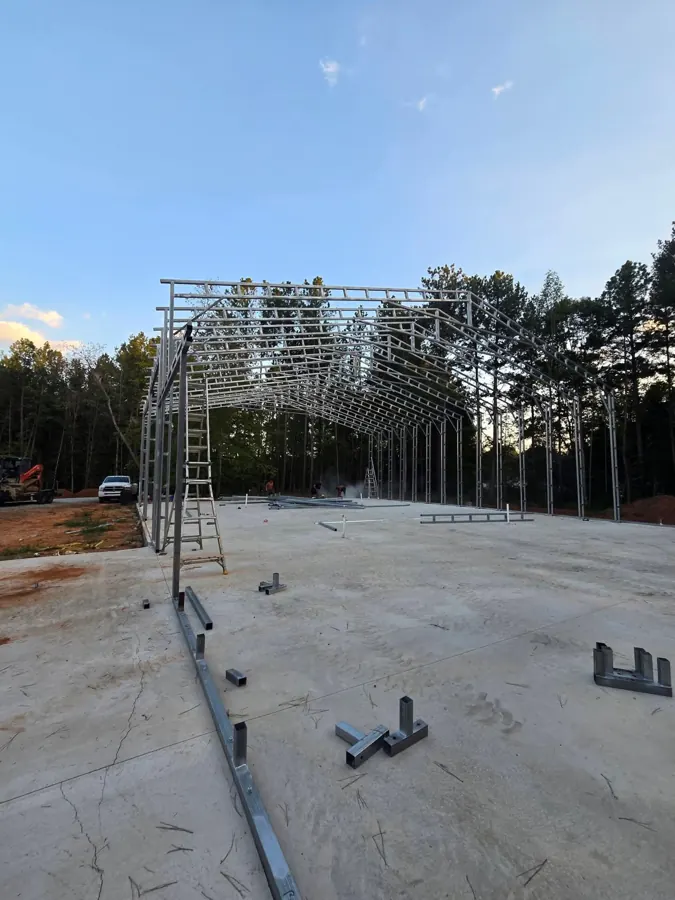 Partly erected steel frame of a 50 by 80 metal building on a concrete slab, with no roof or wall panels, loose steel parts on the slab and a crack in the foreground